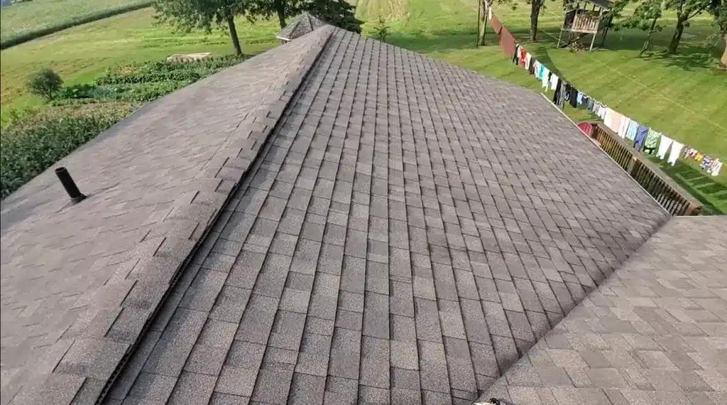 Gray shingled farmhouse roof with ridge, surrounded by trees, grass, and laundry hanging nearby.