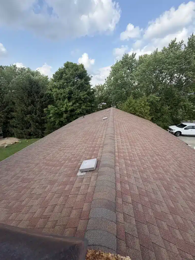 Brown shingled roof ridge with vents in Baden, Ontario, bordered by trees and a white car nearby.