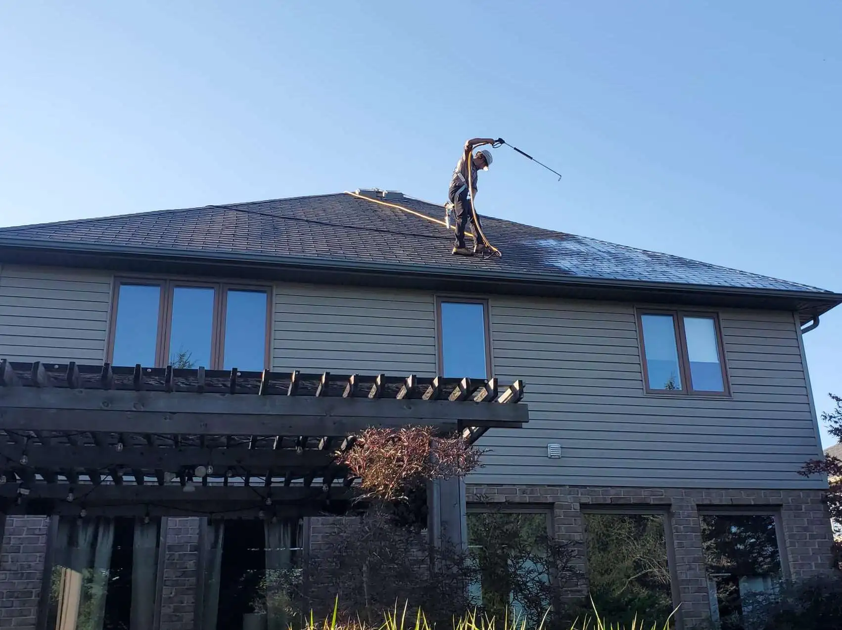 Worker on roof uses spray wand to clean shingles during eco-friendly roof rejuvenation process.