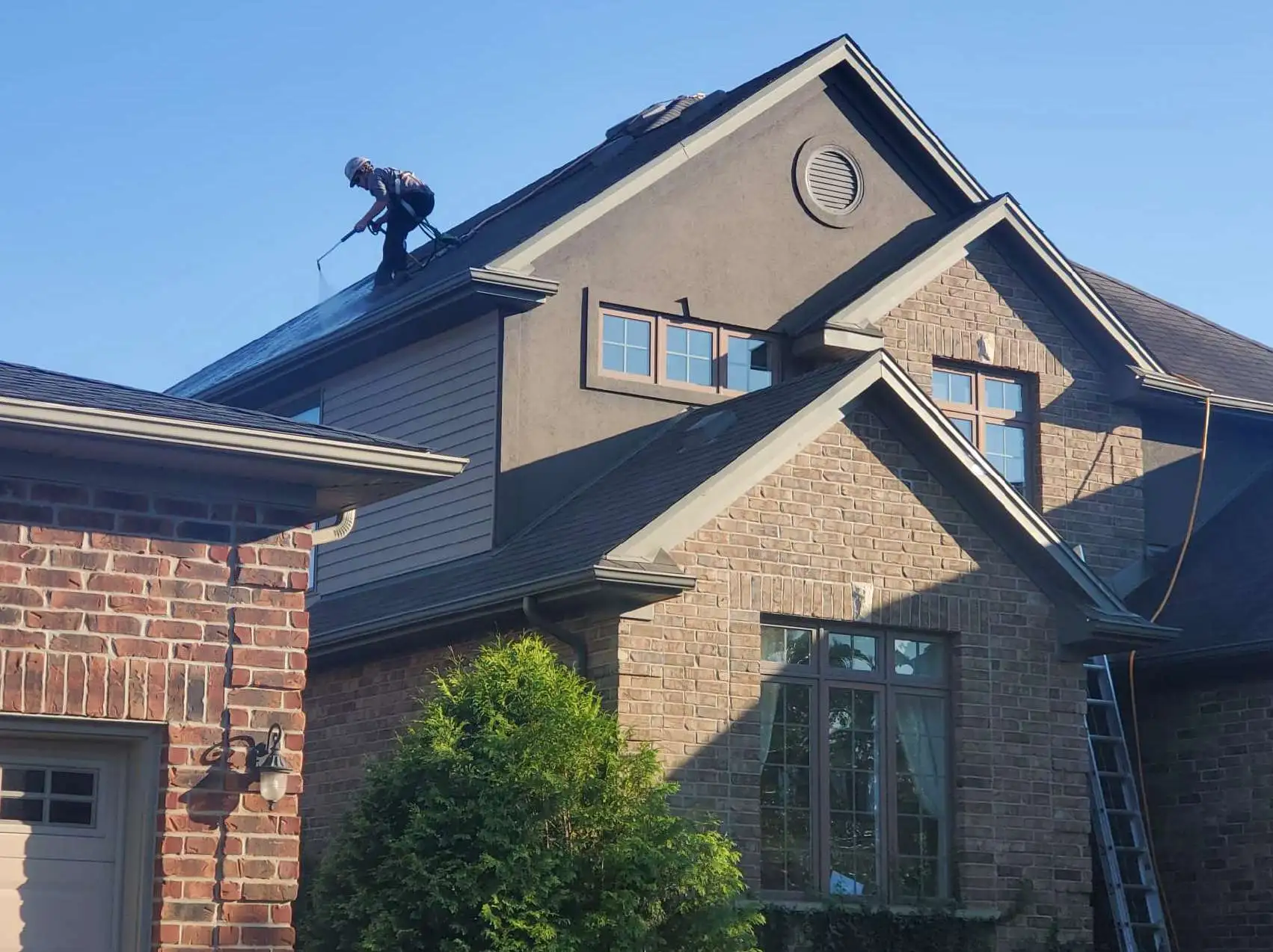 Worker in safety gear applies eco-friendly roof treatment to a two-story brick house.
