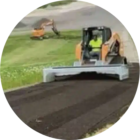 Worker operating compact vehicle spreads soil along roadside for asphalt rejuvenation.