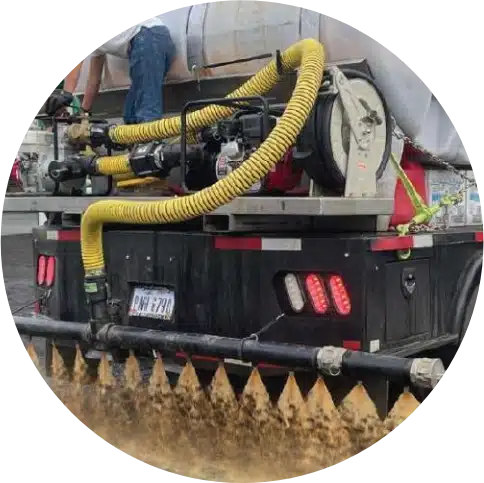 Truck spraying rejuvenation liquid on asphalt, yellow hoses attached above rear nozzles.