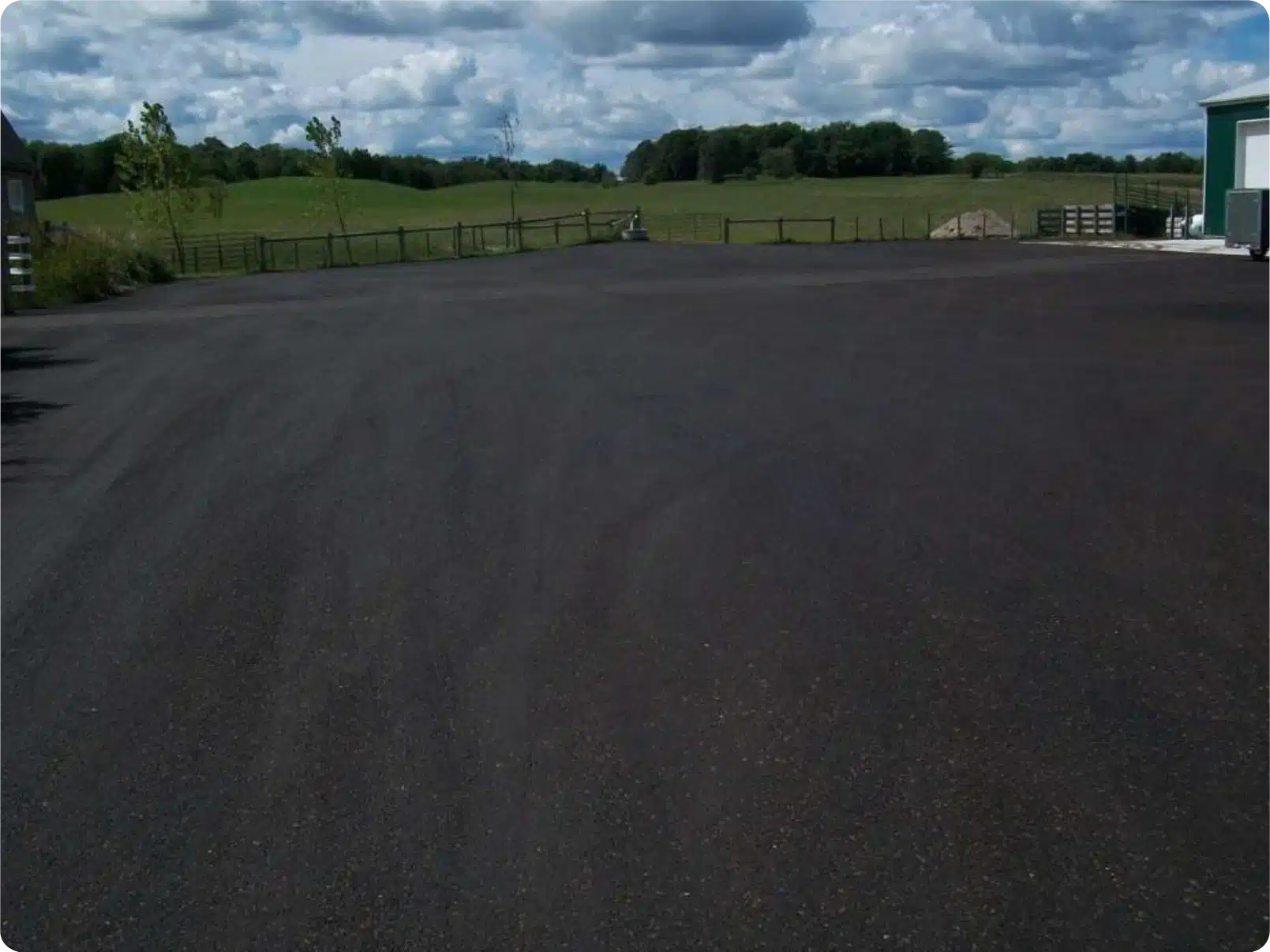 Recycled asphalt parking lot, fenced, bordered by green fields under a cloudy sky.