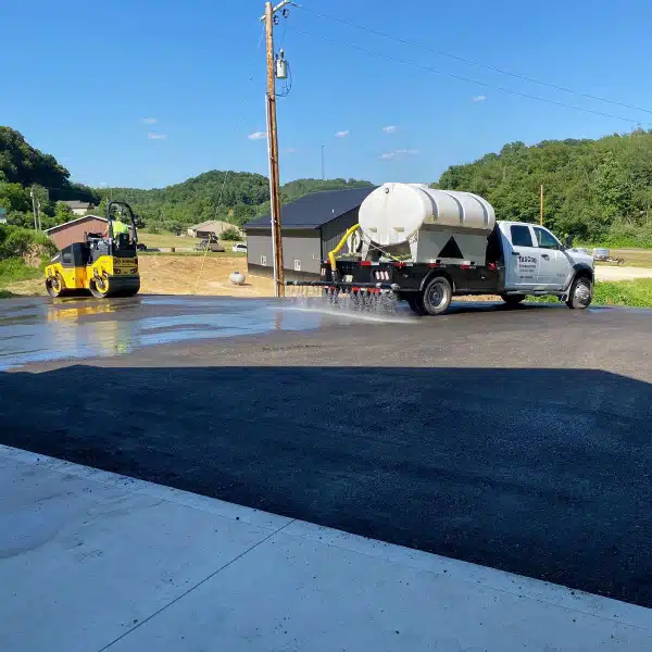 New Project (21) Water truck spraying asphalt beside a steamroller during pavement rejuvenation in a rural area.