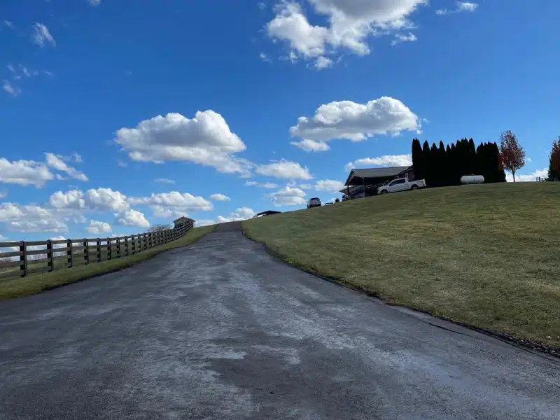 New Project (26) Uphill paved driveway with fresh asphalt leads to house and parked cars beneath clear blue sky.