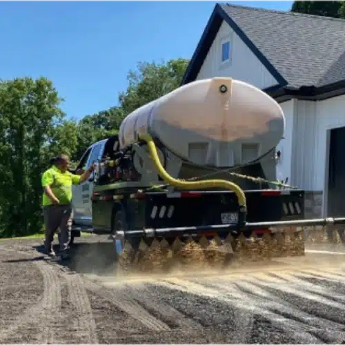 New Project (7) Reactiv8 truck spraying liquid from a large tank onto a gravel driveway with worker operating controls.