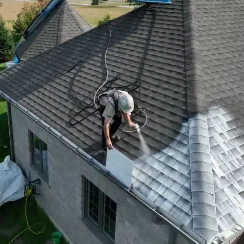 spray Worker in safety gear power washes dark shingles on a sloped roof.